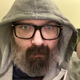 Mike Fahey of Kotaku looks at the camera while wearing a grey hoodie and black rimmed glasses.